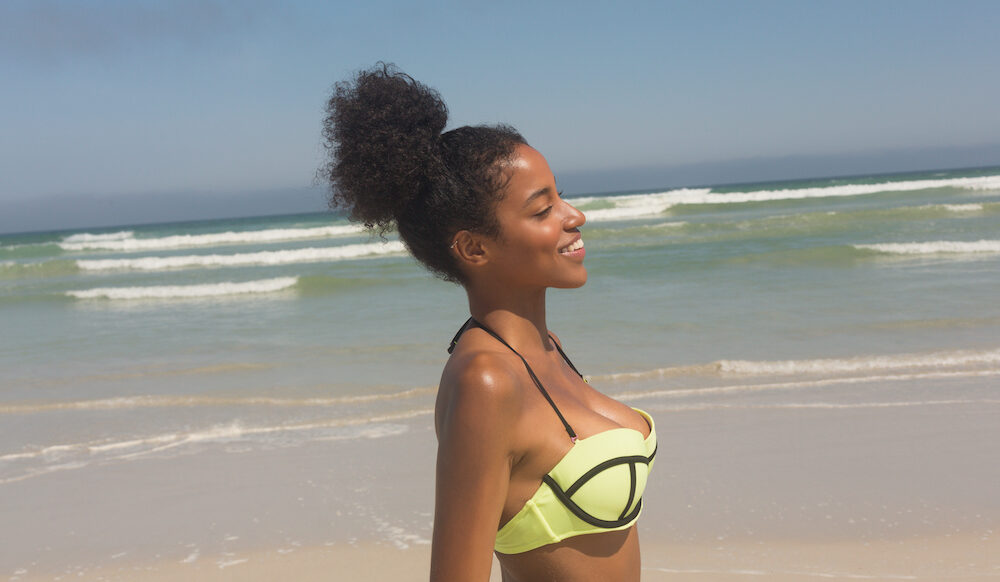 Side view of pretty happy young African American woman in green bikini standing on the beach. She is smiling